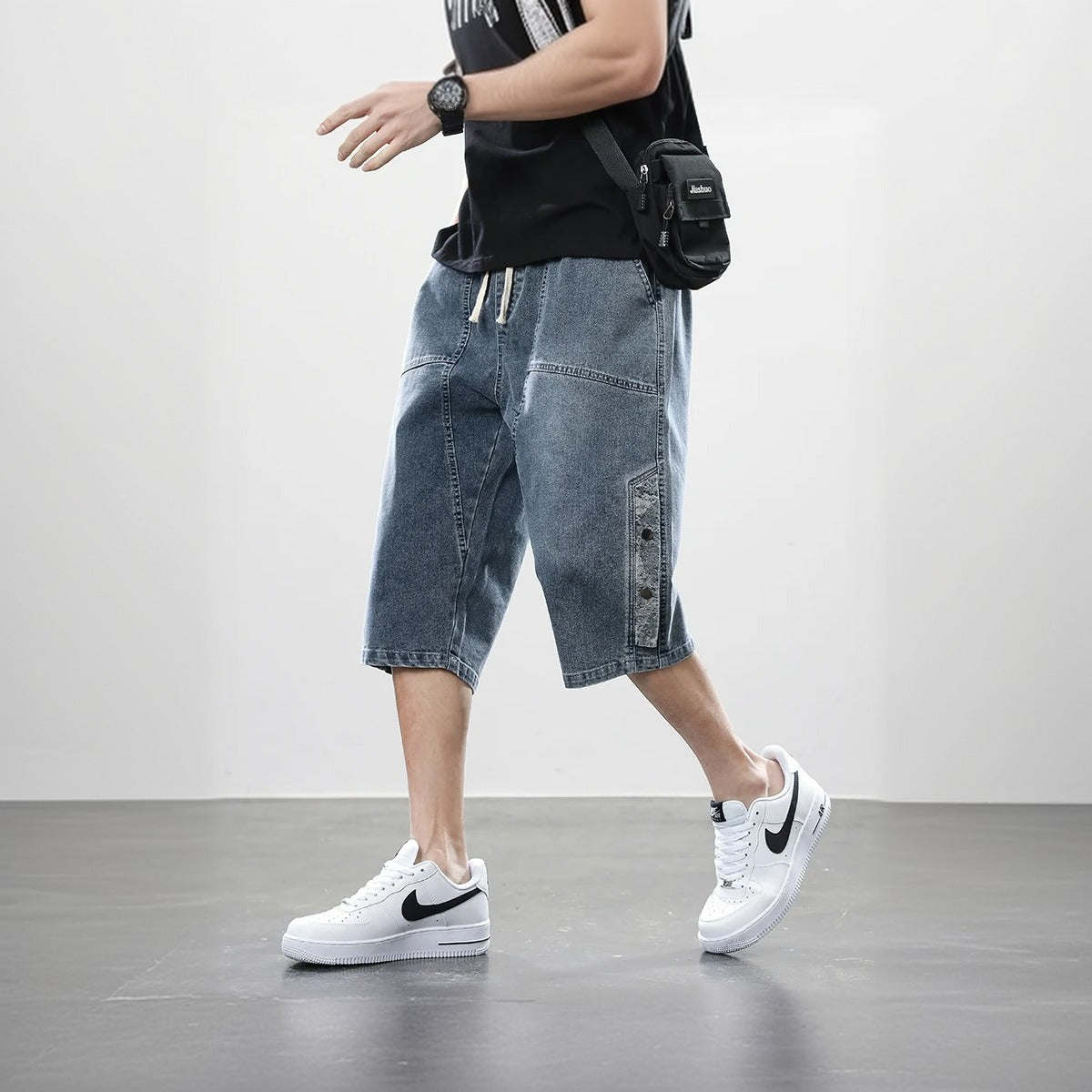  A man walking with baggy denim over the knees blue jeans shorts, a black shirt and wearing a pair of white sneakers.