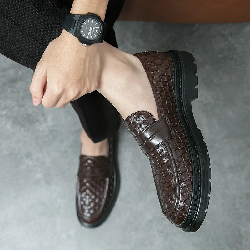 A man with legs folded wearing a pair of brown woven loafers, with black soles , no socks, a black watch and a pair of black corduroy pants.