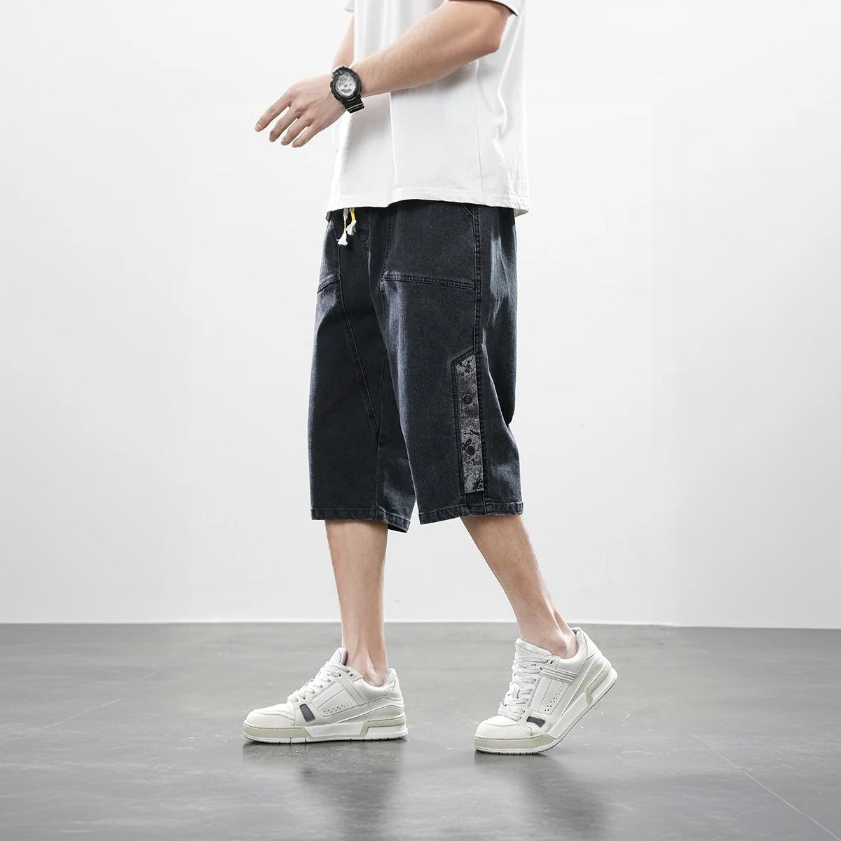 A man is walking, wearing baggy denim knee-length black jean shorts, a white shirt, and white sneakers.